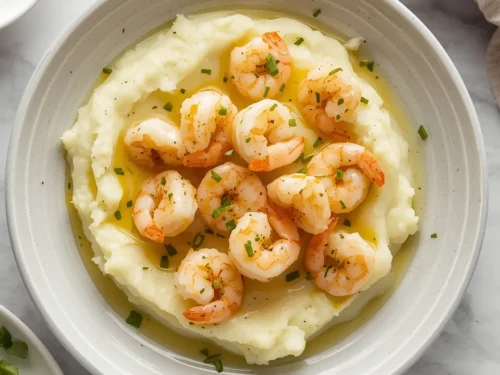 A serving of garlic butter shrimp plated over fluffy mashed potatoes, garnished with herbs.